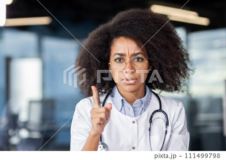 Serious african american doctor in white lab coat raising index finger with rigorous facial expression. Experienced cardiologist emphasising harm of smoking to cardiovascular system for adults. Serious african american doctor in white lab coat raising index finger with rigorous facial expression. Experienced cardiologist emphasising harm of smoking to cardiovascular system for adults. 111499798