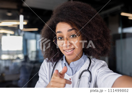 Diverse medic student in eyeglasses with bushy hairdo pointing at camera on blurred background. Pretty female intern recording video about importance of regular body checks for disease preventing. Diverse medic student in eyeglasses with bushy hairdo pointing at camera on blurred background. Pretty female intern recording video about importance of regular body checks for disease preventing. 111499802