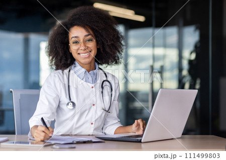 Friendly latino paediatrician in medical coat sitting by desktop with laptop and making notes by hand indoors. Happy brunette lady feeling satisfied with occupation and possibility of healing kids. Friendly latino paediatrician in medical coat sitting by desktop with laptop and making notes by hand indoors. Happy brunette lady feeling satisfied with occupation and possibility of healing kids. 111499803