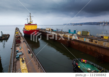 Red icebreaker docked in shipyard floating dry dock for repair, under cloudy sky. Maritime industrial marine vessel maintenance, refit process underway. Heavy steel ship stability ensured. 111500842