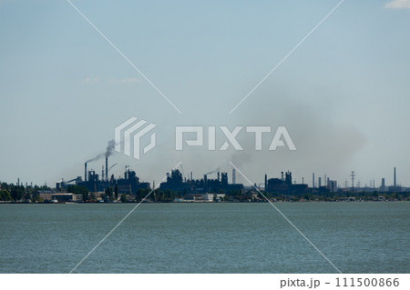 Industrial skyline dominated by metallurgical plant emissions, smoke billowing against sky, symbolizing eco pollution issues, with water foreground. 111500866