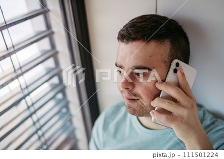 Young man with Down syndrome sitting by window, making phonecall with smartphone. 111501224