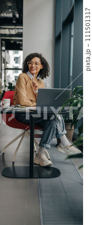 Smiling female manager in eyeglasses working on laptop while sitting in modern office near window 111501317
