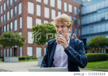 Businessman, freelancer or manager working outdoors in city park. Man with earbuds drinking coffee and listening music. Concept of working remotely. 111501828