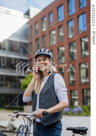 Beautiful middle-aged woman commuting through the city by bike, phone calling. Female city commuter traveling from work by bike after long workday. 111501829