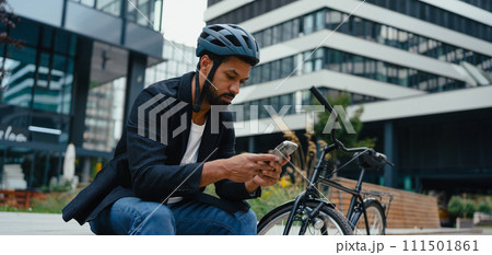 City commuter, sitting on bench, scrolling on smartphone. Businessman traveling from work to home on bike. City lifestyle. 111501861