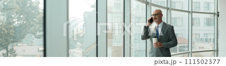 Header of confident mature male solopreneur in grey suit speaking on smartphone while looking through large window in office center 111502377