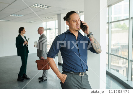 Young Asian male solopreneur in shirt and pants speaking on mobile phone while standing in front of camera in office center 111502473