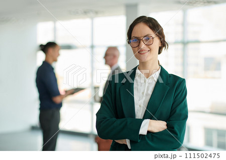 Young successful female chief executive officer in eyeglasses and formalwear looking at camera with smile against two colleagues 111502475