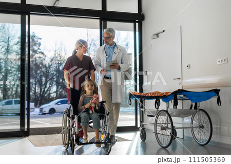 Friendly pediatrician nurse talking, pushing little patient in wheelchair. Cute preschool girl trusting her doctor and nurse in hospital. Friendly pediatrician nurse talking, pushing little patient in wheelchair. Cute preschool girl trusting her doctor and nurse in hospital. 111505369