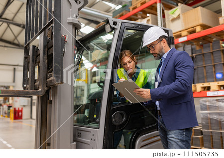 Female forklift driver talking with manager in warehouse, order picking. Warehouse worker preparing products for shipmennt, delivery, checking stock in warehouse. 111505735