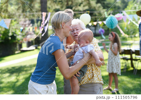 Great-grandmother holding crying little baby in her arms. Family summer garden party. 111505796