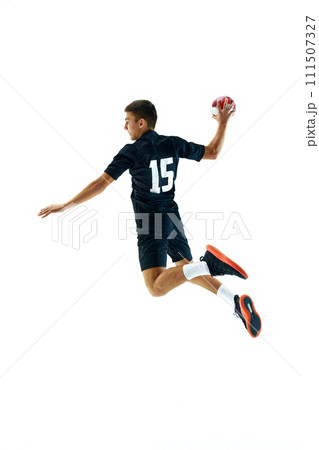 Full-length side view image of young man in dark uniform, handball player in motion, in a jump with ball during game, training against white studio background 111507327