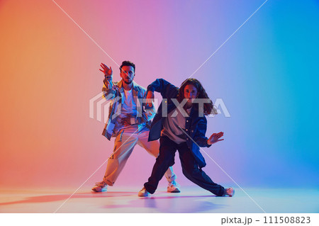 Dynamic lifestyle. Man and woman, hip-hop dance moves sync in motion against gradient studio background in neon light. Dynamic lifestyle. Man and woman, hip-hop dance moves sync in motion against gradient studio background in neon light. 111508823