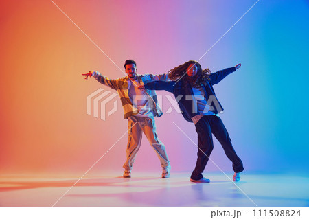 Dynamic photo of two fashion dressed people, dancing in motion against gradient studio background in neon light, filter. 111508824