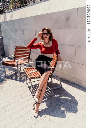 Portrait of a woman on the street. An attractive woman in glasses, a red blouse and a black skirt is sitting on a bench outside. 111509042