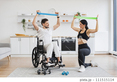 Wife helping mature man in wheelchair lifting arms with stretching bands. 111515114