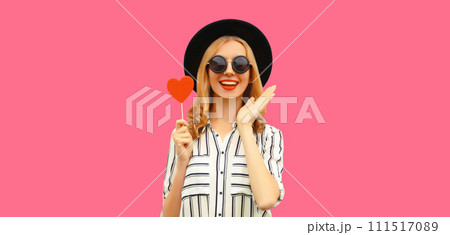 Portrait of happy cheerful young woman with sweet red heart shaped lollipop on stick on pink 111517089