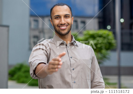 Portrait of a successful and smiling young businessman, hispanic man standing outside an office building and extending his hand to the camera in greeting. Portrait of a successful and smiling young businessman, hispanic man standing outside an office building and extending his hand to the camera in greeting. 111518172