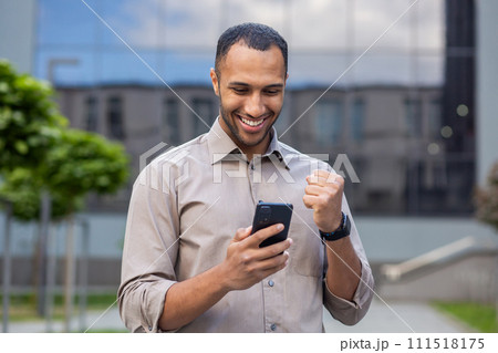 Young muslim male businessman standing on street near office building and using mobile phone, looking at screen and happy with received news and message showing victory gesture with hand. Young muslim male businessman standing on street near office building and using mobile phone, looking at screen and happy with received news and message showing victory gesture with hand. 111518175