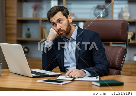 A young businessman man in a suit sits in the office at a desk in front of a laptop, is sad and upset, leaning his head and hand. He looks thoughtfully to the side. A young businessman man in a suit sits in the office at a desk in front of a laptop, is sad and upset, leaning his head and hand. He looks thoughtfully to the side. 111518192