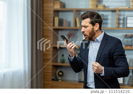Angry young male businessman standing in the office in a suit and emotionally talking on the phone. Holds a mobile phone and shouts into the loudspeaker, solves questions and problems. Angry young male businessman standing in the office in a suit and emotionally talking on the phone. Holds a mobile phone and shouts into the loudspeaker, solves questions and problems. 111518210