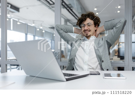 A happy young man businessman sits in the office at the table and smilingly looks at the laptop screen, satisfied with the work done, rests with his hands behind his head. 111518220
