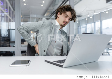 Tired young man businessman sitting at the desk in the office, working on a laptop and holding his hand behind his back, taking a break, feeling pain and tension. 111518221