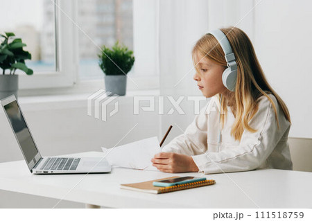 Happy Caucasian girl studying at home using a laptop for elearning and online games, with headphones on and a notebook on the table The modern digital connection provides a virtual school experience, 111518759