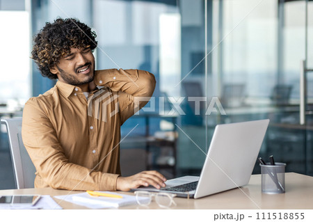 A young Indian man is suffering from severe neck pain. Tired man sits at the desk in the office and massages his hand. 111518855