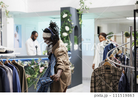 Cheerful african american man in headphones holding shirt on hanger while shopping in clothing store. Customer in good mood listening to music while choosing new outfit in fashion boutique 111519863