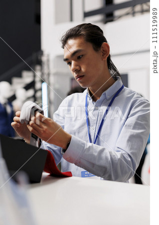 Asian manager folding fashionable ties in modern boutique, working at store visual. Cheerful stylish employee arranging new fashion collection, checking clothes price in shopping centre 111519989