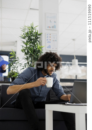 Arab company employee drinking coffee and reading customer email on laptop in office. Business start up focused entrepreneur holding tea mug and managing project in coworking space Arab company employee drinking coffee and reading customer email on laptop in office. Business start up focused entrepreneur holding tea mug and managing project in coworking space 111519990