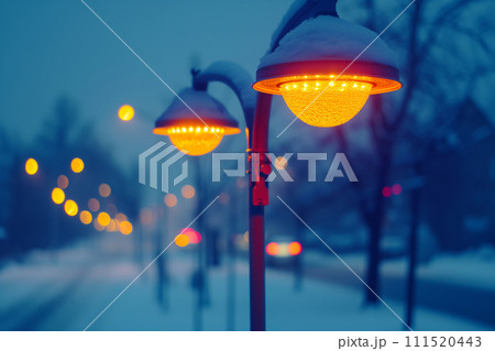 Glowing street lamps line a snowy avenue at dusk, casting a warm amber light over the quiet winter landscape. 111520443