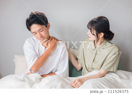 朝寝室で肩こりに悩む男性と心配する女性・夫婦・カップル 111526375