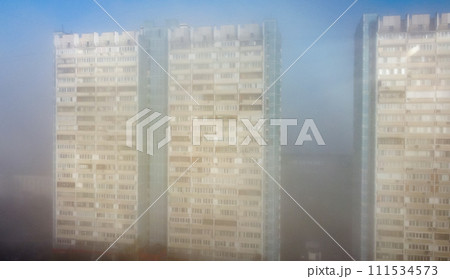 A white-and-blue multi-storey residential building is shrouded in fog in the early spring morning. 111534573