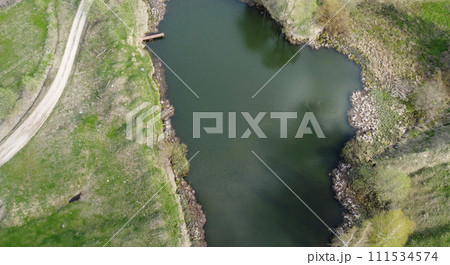 A dirt road near a village pond in the middle of a green meadow. Aerial view. 111534574