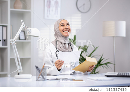 Smiling female Muslim doctor in a hijab and white coat holding papers in a modern clinic office, showcasing professional healthcare. Smiling female Muslim doctor in a hijab and white coat holding papers in a modern clinic office, showcasing professional healthcare. 111537456