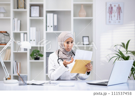 A focused Muslim female doctor in a hijab and white coat examines documents in her medical office, symbolizing healthcare diversity. A focused Muslim female doctor in a hijab and white coat examines documents in her medical office, symbolizing healthcare diversity. 111537458