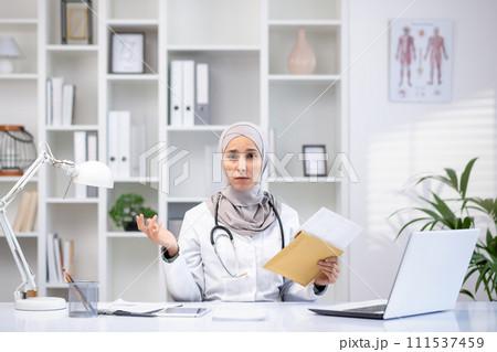 Professional woman in hijab and lab coat in a clinic office, engaging in patient consultation with laptop and medical equipment. Professional woman in hijab and lab coat in a clinic office, engaging in patient consultation with laptop and medical equipment. 111537459