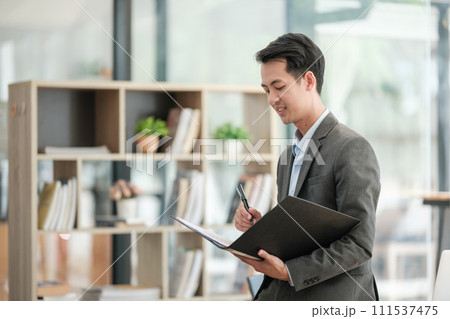 Smiling young bearded business man standing near desk in light office background. Achievement business career concept. Smiling young bearded business man standing near desk in light office background. Achievement business career concept. 111537475