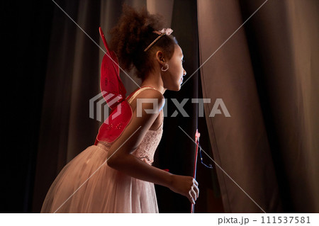 Side view young Black girl wearing fairy costume with wings peeking over curtain backstage in theater copy space 111537581