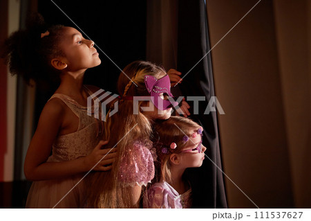 Side view portrait of three little girls wearing costumes peeking over curtain backstage in theater copy space 111537627