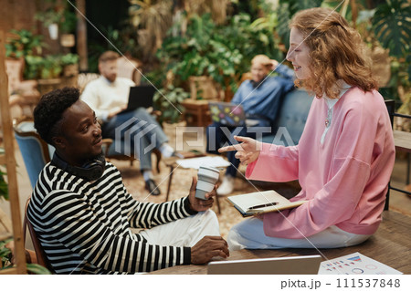 Side view portrait of young woman wearing pink talking to colleague in coworking room and smiling 111537848
