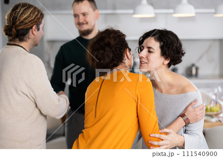 Women hug each other and kiss on the cheek while greeting each other before a festive dinner. Men shaking hands in background 111540900