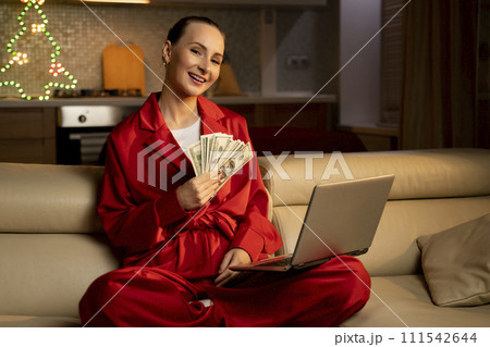 Elated Woman in Red Pajamas Holding Cash While Using Laptop Indoors During Christmas Season Elated Woman in Red Pajamas Holding Cash While Using Laptop Indoors During Christmas Season 111542644