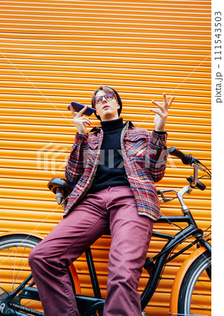 Young hipster man recording voice message on phone while sitting on cruiser bicycle near orange wall. Student using 5g network. Eco friendly transport for carbon footprint. Sustainable lifestyle. 111543503