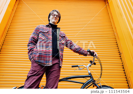 Bottom view Young stylish hipster man standing with a cruiser city Bicycle near orange Wall. Student Resting after ride bicycle. Eco friendly transportation for carbon footprint. Sustainable lifestyle Bottom view Young stylish hipster man standing with a cruiser city Bicycle near orange Wall. Student Resting after ride bicycle. Eco friendly transportation for carbon footprint. Sustainable lifestyle 111543504