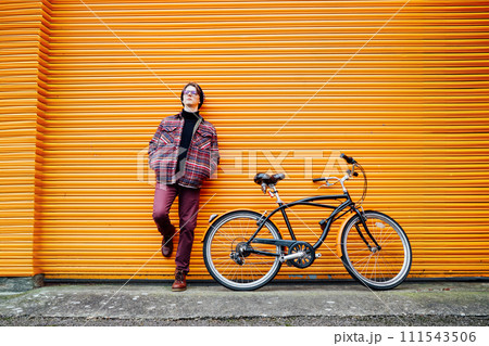 Young stylish hipster man standing with a cruiser city Bicycle near orange Wall. Student Resting after ride bicycle. Eco friendly transportation for carbon footprint. Sustainable lifestyle 111543506