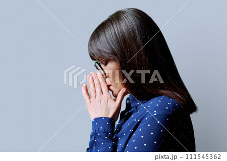 Profile view of mature sad woman, with closed eyes, folded hands, gray background 111545362